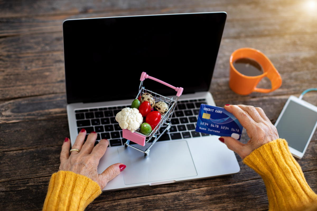 senior woman at a laptop with a credit card and tiny shopping cart to suggest online shopping and delivery