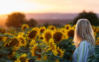 a senior woman stands in a field of sunflowers at sunset.